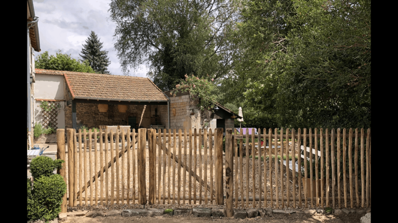 Dubbele franse rondhoutpoort van 120 cm breed bij 120 cm hoog per poortdeel bevestigd aan hekwerk van kastanjehouten palen en halfronde rasterlatten als doorgang naar de oprit bij een huis.