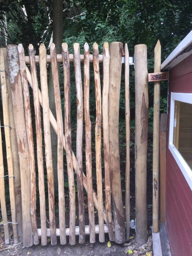 Franse rondhoutpoort van 150 cm hoog en 80 cm breed als doorgang in een tuin tussen schapenhek en een schuurtje.