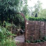 Wilgentenen toegangsboog vastgezet aan een wilgentenen scherm dat in een boog loopt. De wilgentenen staan in de tuin waar veel groen en bomen zijn. Scherm wordt gebruikt als afrastering van een deel in de tuin.