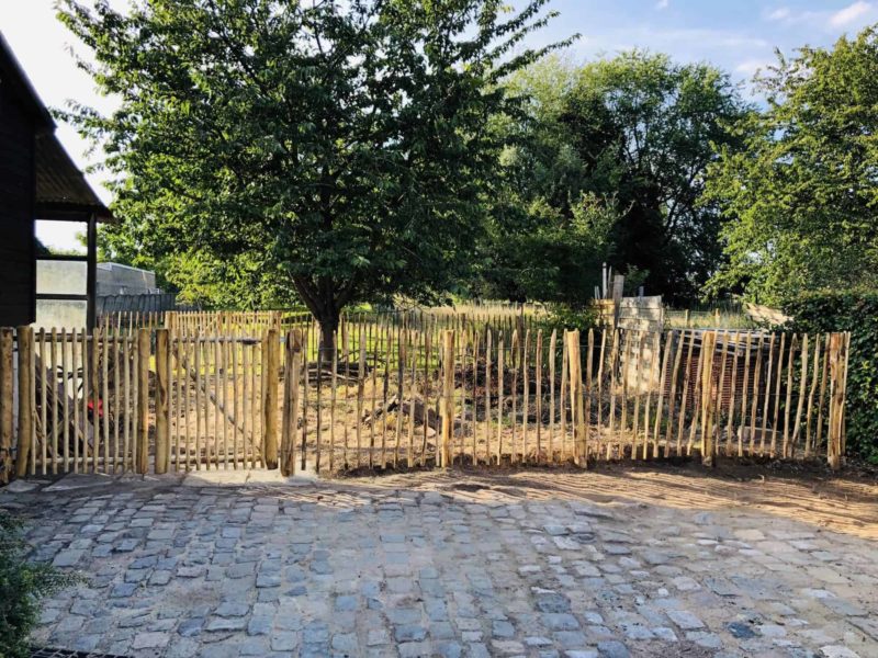 Dubbele Franse rondhoutpoort van 120 cm hoog en 100 cm breed per poortdeel als doorgang naar de tuin met een afscheiding van schapenhek van 120 cm hoog en een latafstand van 8 cm