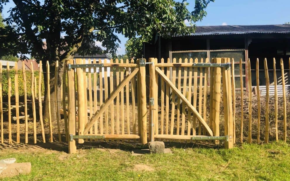 Dubbele Franse rondhoutpoort van 120 cm hoog en 100 cm breed per poortdeel als doorgang naar de tuin met een afscheiding van schapenhek van 120 cm hoog en een latafstand van 8 cm