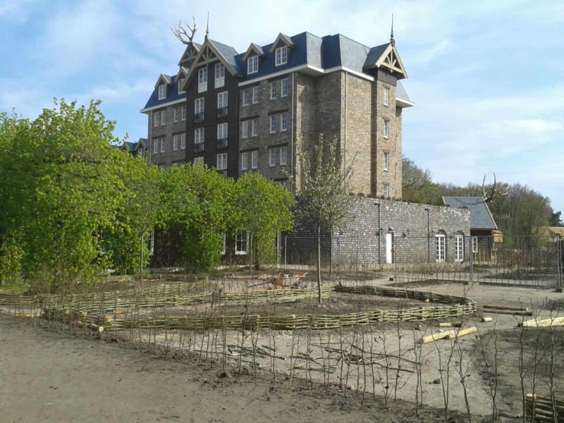 Wilgentenen schutting gevlochten tussen twee heggen en onder een hele grote boom. Er is veel groen te zien en de wilgentenen gaan mooi op in al het groen van de tuin.