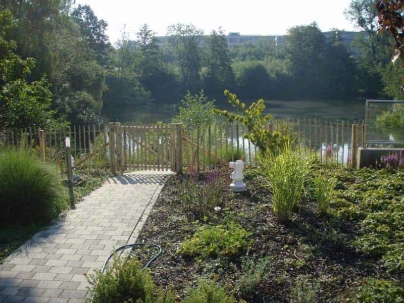 Franse rondhoutpoort van 100 cm hoog als doorgang bij een omheining in een tuin bij het water. De omheining is van schapenhek in de maat 100 cm met een latafstand van 8 cm