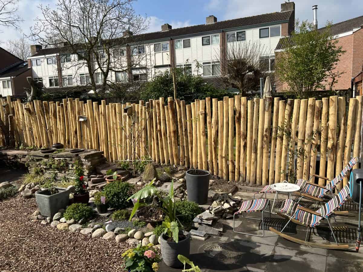 Op de foto is een alternatieve manier te zien van afrastering in de tuin. Hier is gebruik gemaakt van kastanjehouten palen met een diameter van 10 - 12 cm en een lengte van 250 cm. Deze kastanjehouten palen zijn naast elkaar bevestigd in een rechte rij en fungeren zo als afrastering in de tuin. Naast de palenrij staan aan de rechter kant twee schommelstoelen met zomerse kleuren en een wit tafeltje in het midden. Aan de andere kant van de foto ziet men een tuin die in aanleg is. Hier en daar wat planten en stenen. Om de afratering nog wat te personaliseren zijn er een paar vogelhuisjes bevestigd aan de palen. Dit geeft een persoonlijke touch en een vrolijke en rustieke uitstraling aan de tuin.