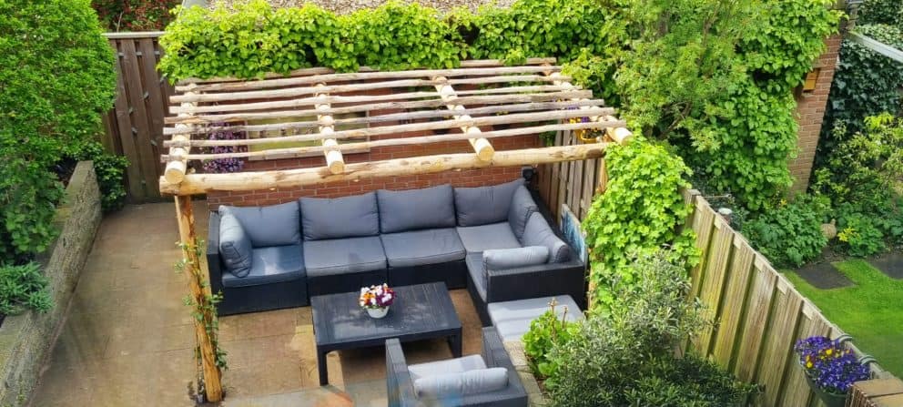 Op de foto is een bovenaanzicht van een kastanjehouten pergola te zien met een mooie natuurlijke lichtbruine kleur. De ronde palen die gebruikt zijn voor de pergola hebben een diameter van 10 - 12 cm en de halfronde palen hebben een diameter van 7 - 9 cm. Onder de pergola staat een zwarte loungeset met grijze kussen. De loungeset bestaat uit een bank, een stoel en een tafel. Op de tafel staat een pot met gekleurde bloemen. De pergola staat in de tuin tegen een klein schuurtje aan. Rondom de pergola is veel groen te zien. De pergola heeft klimop begroeing aan de pilaren vanaf de grond. Dit zorgt voor een intieme en natuurlijke uitstraling.