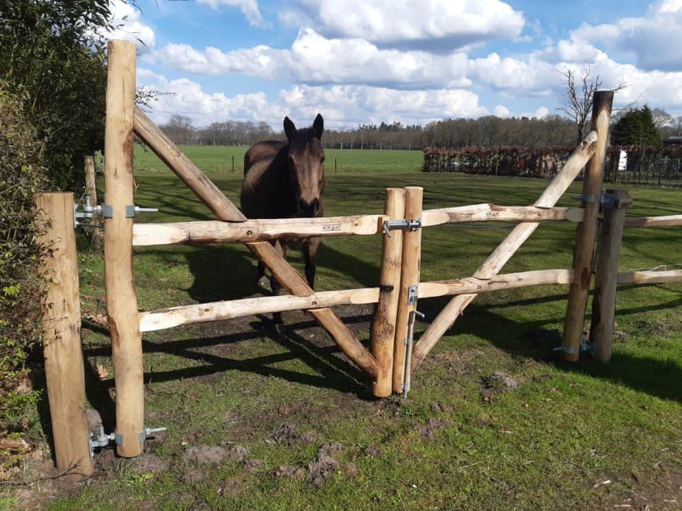 Op de foto is een weiland met een bruin paard te zien. Het paard staat voor de post en rail poort. De post en railpoort is een dubbele 2 niveau poort met trekboom met een breedte van 150 cm en een hoogte van 120 cm per poortdeel. Deze poort is gebruikt als afrastering en toegang voor een paardenwei. De veersluiting zit aan de voorkant van de poort en onderaan aan de rechterkant is een grondpen bevestigd. Naast de post en rail toegangspoort staat post en rail hekwerk. Dit hekwerk is ook een niveau 2 post en rail. De lichte en wat donkere kleuren bruin van het hout gaan goed op in de omgeving, zo geeft dit een rustieke en landelijke sfeer en daarnaast zit het paard veilig in de wei.