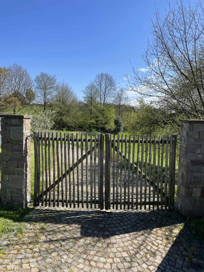 Op de foto is een gesloten rustieke dubbele Franse rondhoutpoort van 150 cm hoog en 138 cm breed van Adequat te zien. De poort is bevestigd aan twee stenen pilaren van ongeveer 200 cm hoog. Op de foto is de oprijlaan te zien, deze bestaat uit klinkers. De foto is genomen vanaf de kant van het woonerf en kijkt uit op de Duitse groene natuur waarin veel groen en bomen te zien zijn. De poort voegt een rustieke en landelijke sfeer toe aan de foto en gaat goed op in de natuur van de omgeving.