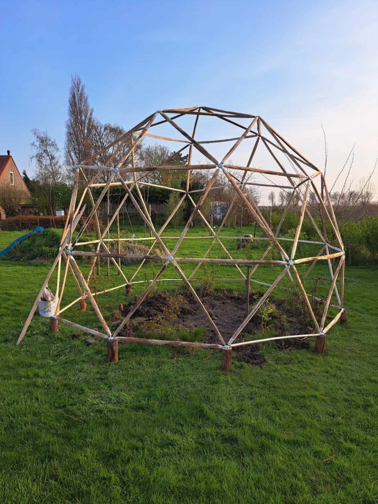 Een bolvormig bouwwerk in het weiland, gemaakt van kastanjehouten latten. Deze geodome staat boven een moestuin en zal het gewas beschermen tegen vogels