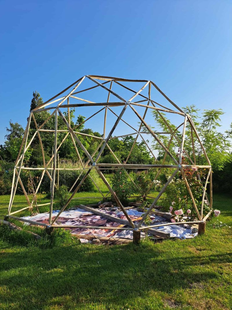 Bolvormige contructie van kastanjehout, geplaatst over een bloementuin. De geodome staat in een tuin op het gras en er staat een houten schommel naast