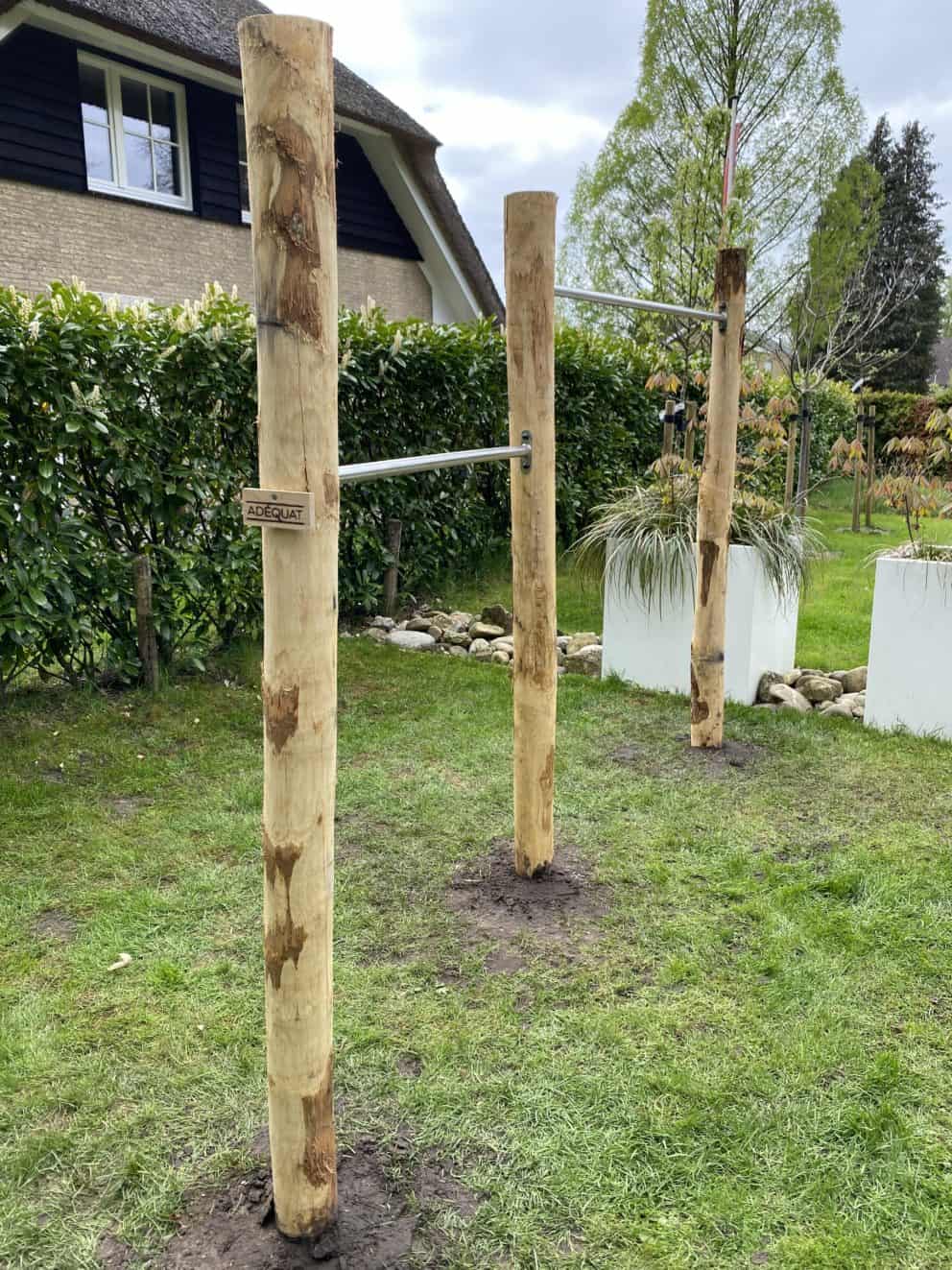 Op de foto staat een duikelrek voor kinderen van kastanje palen in de diameter 10-12 cm. Het duikelrek heeft twee verschillende niveaus en staat in de tuin. Er is gebruik gemaakt van drie kastanjehouten palen in dezelfde lengte. Op de achtergrond is een stuk heg, een deel van een woning en twee witte planten bakken zichtbaar.