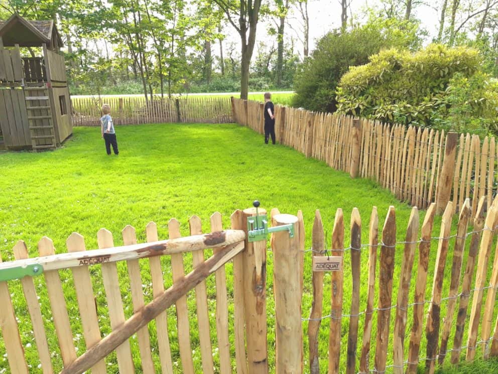 Frans schapenhek van 100 cm hoog met een latafstand van 4 cm in combinatie met een rondhoutpoort van 150 cm breed en 100 cm hoog met een simpele sluiting. De poort sluiting zit aan de buitenkant van de poort. In de omheining staan twee kinderen van het kinderdagverblijf. Ze staan met hun rug naar de camera en geven een duimpje omhoog.