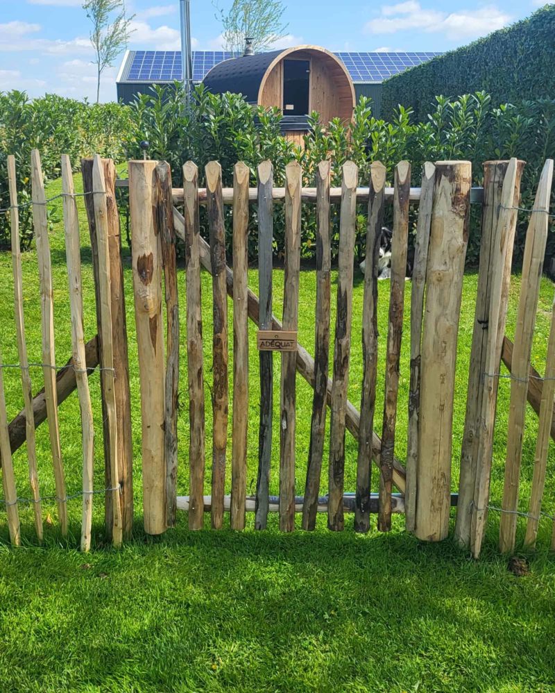 Schapenhekwerk van 120 cm hoog met een latafstand van 4 cm in combinatie met een franse rondhoutpoort van 100 cm breed en 120 cm hoog met een simpel slot bevestigd aan de binnenkant van de poort. Deze producten worden gebruikt als afrastering van een deel van de tuin. Achteraan op de foto is een ronde sauna te zien.