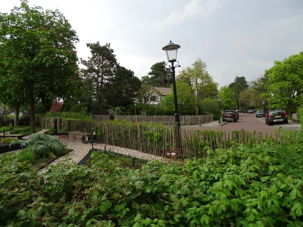 Frans schapenhek van 100 cm hoog met latafstand 4 cm gebruikt als omheining voor een bloementuin. De foto is genomen vanaf een struik en kijkt uit op de bloementui die al een paar bloemen in bloei heeft staan. Op de grond is een wit kiezelstenen pad te zien en aan de linker kant staan twee houten bankjes.