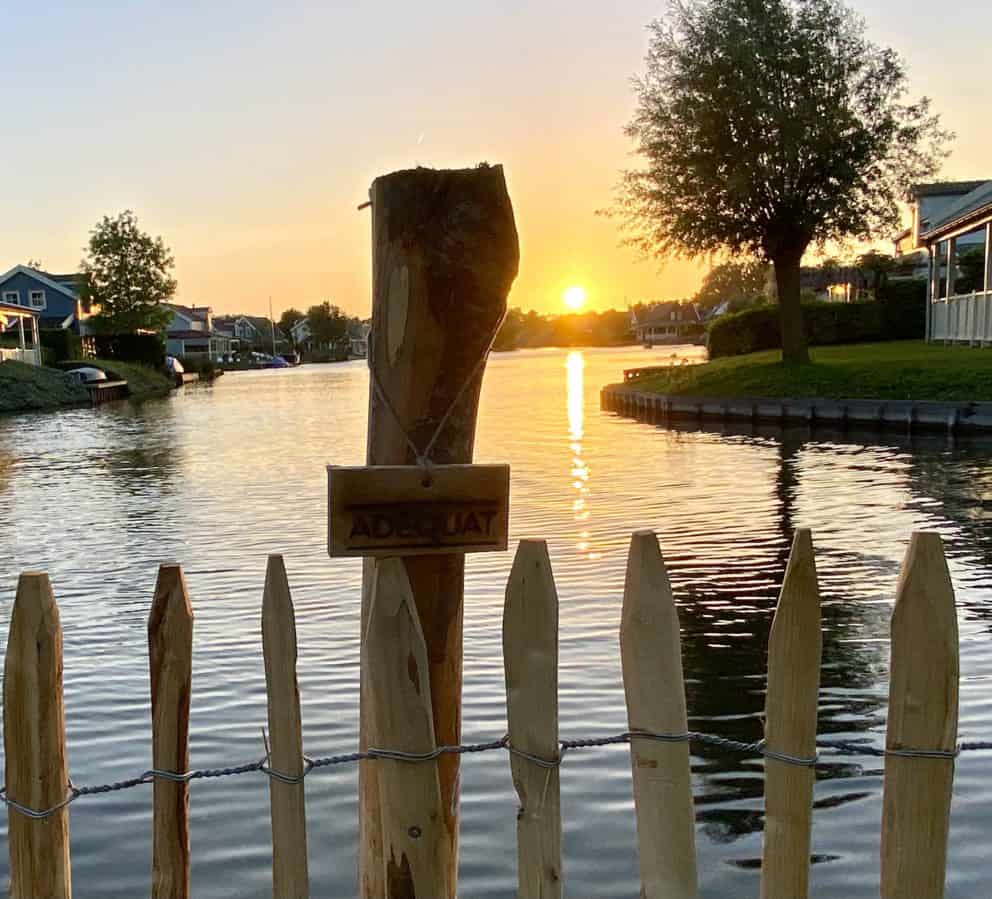 Een close up van frans schapenhek van 80 cm hoog met een latafstand van 4 cm aan het water met zonsondergang. Op de foto is zowel rechts als links woningen te zien. In het midden van de foto kijk je uit op water.