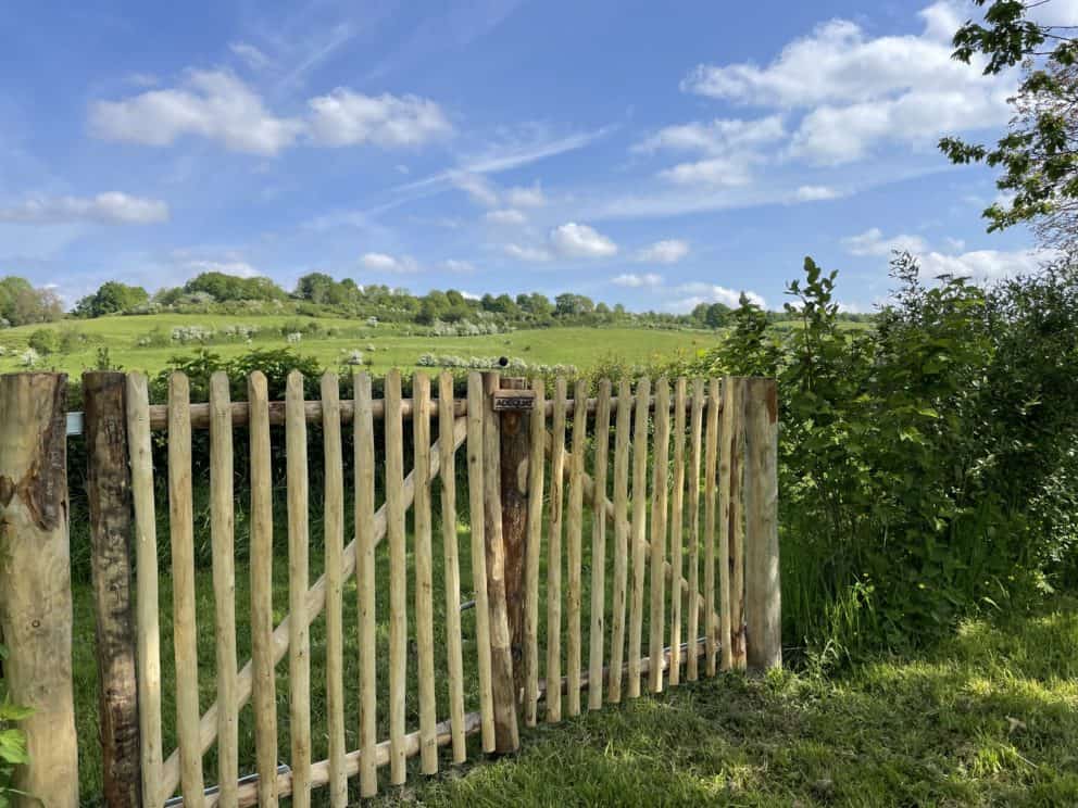 Dubbele franse rondhoutpoort van 100 cm breed en 100 cm hoog met een simpele sluiting aan de buitenkant. De foto is genomen vanaf de voorkant van de poort en je kijkt uit over een mooi groen landschap. De poort zorgt voor een nog landelijkere uitstraling.