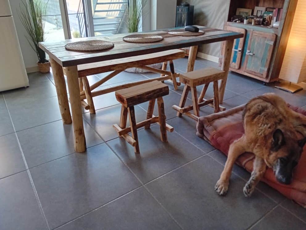 Boomstamkankje van kastanjehout van 150 cm breed en twee kastanjehouten krukjes. Deze staan bij een tafel binnen in de woning. De bankjes en de tafel stralen een Ibiza look uit.