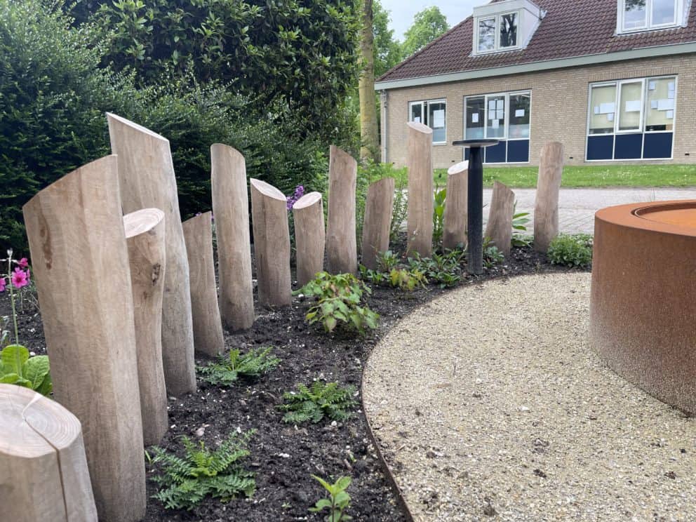 Speelse afscheiding van geschilde en geschuurde robinia palen met een diameter van 12 - 14 cm. De robinia palen zijn in verschillende hoogtes geplaatst in een halve cirkel om zo een creatieve draai te geven aan een pad in de tuin. De palen staan aan de linker kant van de foto en aan de rechter kant is een kiezelpad te zien met daarop een installatie van corten staal.