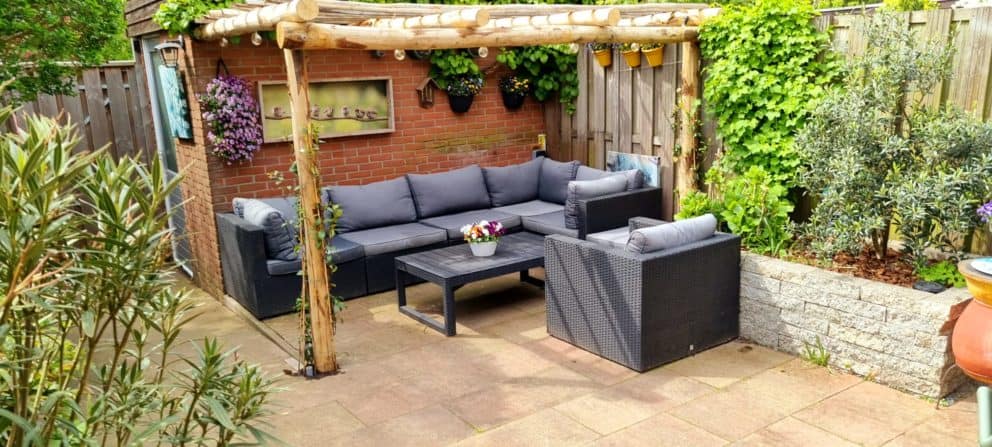 Op de foto is een kastanjehouten pergola te zien met een mooie natuurlijke lichtbruine kleur. De ronde palen die gebruikt zijn voor de pergola hebben een diameter van 10 - 12 cm en de halfronde palen hebben een diameter van 7 - 9 cm. Onder de pergola staat een zwarte loungeset met grijze kussen. De loungeset bestaat uit een bank, een stoel en een tafel. Op de tafel staat een pot met gekleurde bloemen. De pergola staat in de tuin tegen een klein schuurtje aan. Rondom de pergola is veel groen te zien. De pergola heeft klimop begroeing aan de pilaren vanaf de grond. Dit zorgt voor een intieme en natuurlijke uitstraling.