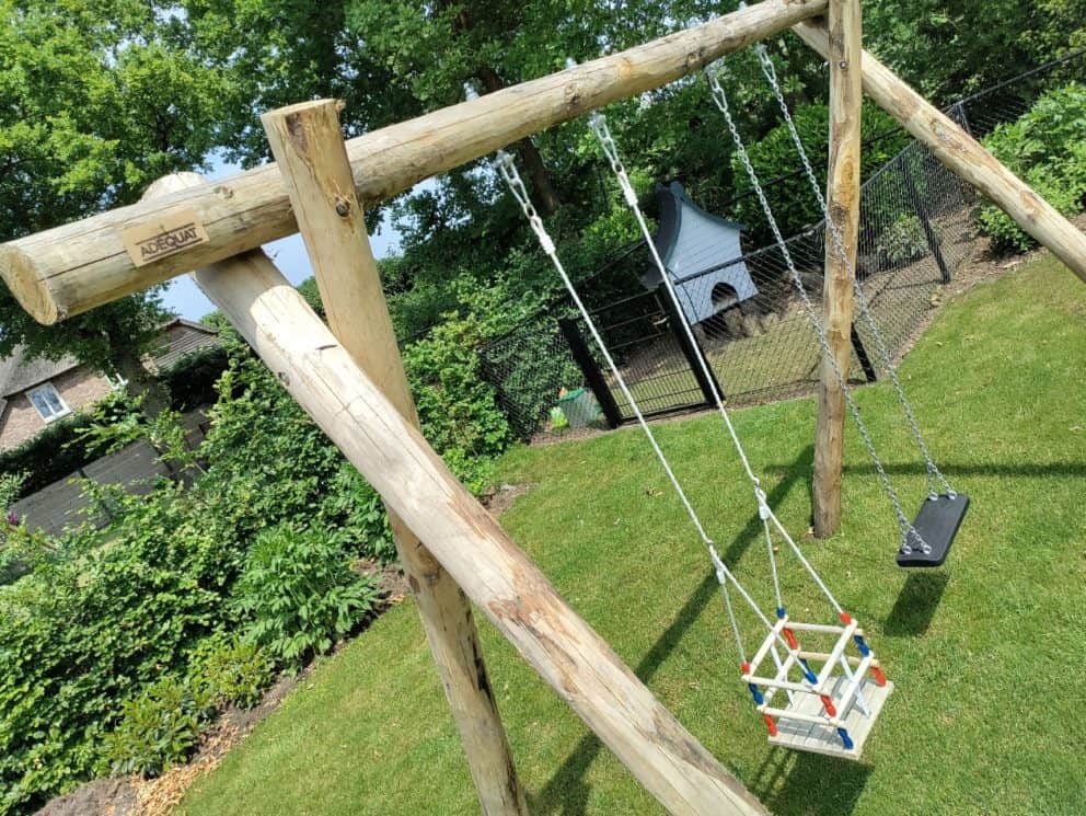 Een dubbele schommel gemaakt van 5 kastanjehouten palen met een diameter van 13 - 16 cm en 350 cm lang en 10 - 12 cm en 200 cm lang. Aan de constructie hangen twee schommels, een daarvan is een schommel voor babys. De schommel staat in de tuin naast een kippenhok.