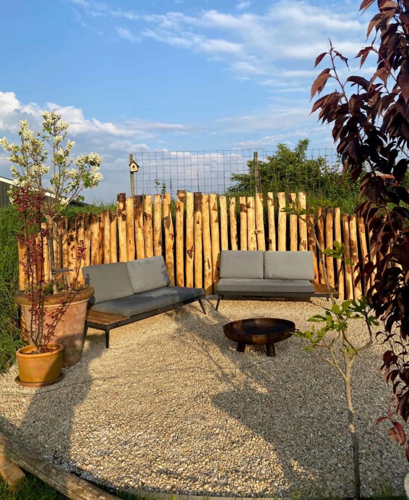 Privacy biedende palenrij van kastanjehouten palen in diverse afmetingen met een diameter van 10-12 cm gebruikt als creatieve afscheiding in de tuin. De palen staan achter twee lounge sets ket gruize kussen. Dit zorgt voor een speelse, maar rustieke uitstraling.